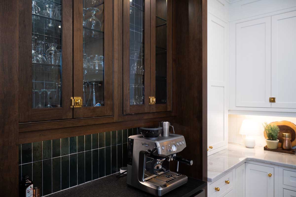 A modern kitchen corner with a stainless steel espresso machine on a black counter, dark wood cabinets with glass doors above, and white cabinets to the side—perfect inspiration for your next Charlotte remodel.